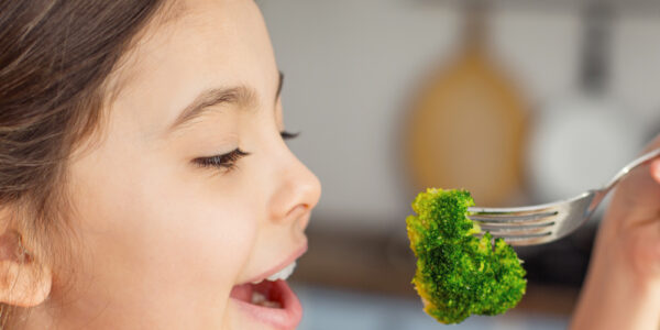 A little girl in a plaid shirt prepares to put a piece of broccoli in her mouth, which she holds on a fork.