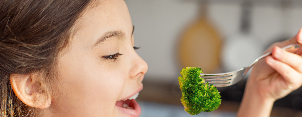 A little girl in a plaid shirt prepares to put a piece of broccoli in her mouth, which she holds on a fork.