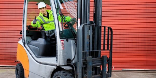 A forklift driver leaning out of his orange-and-gray forklift in front of a red garage door to give the camera a thumbs-up.