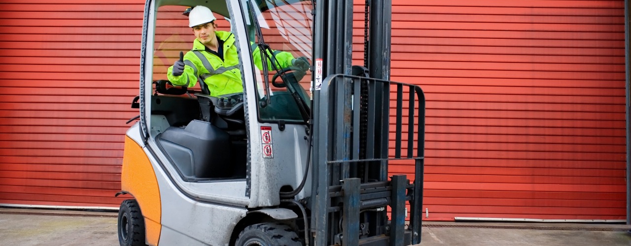 A forklift driver leaning out of his orange-and-gray forklift in front of a red garage door to give the camera a thumbs-up.