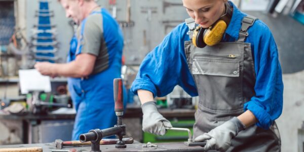 A female metalworker cuts a hole in a piece of metal while a male metalworker stands in the background holding a tool.