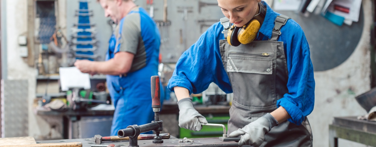 A female metalworker cuts a hole in a piece of metal while a male metalworker stands in the background holding a tool.