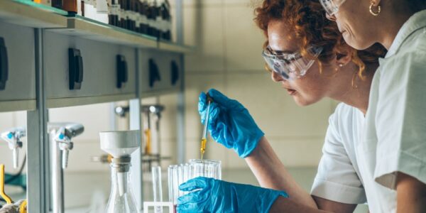 Two female chemistry scientists performing experiments at a lab bench. They wear protective glasses and gloves.