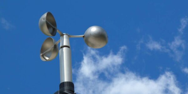 A cup style anemometer on top of a large pole. It has a chrome finish and is not currently spinning.