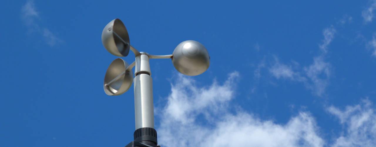 A cup style anemometer on top of a large pole. It has a chrome finish and is not currently spinning.