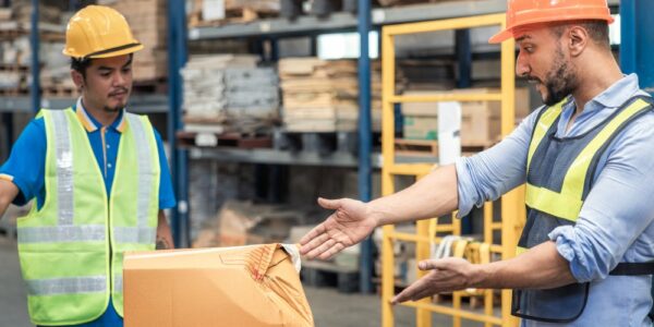 An exasperated worker gesturing to a broken box on a warehouse floor while a forklift operator looks on.