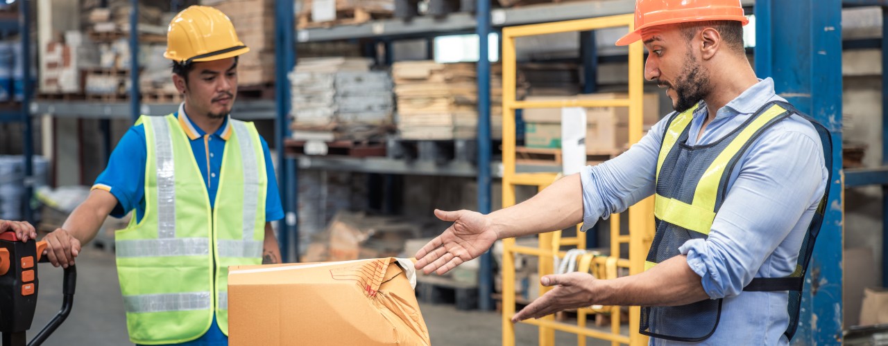 An exasperated worker gesturing to a broken box on a warehouse floor while a forklift operator looks on.