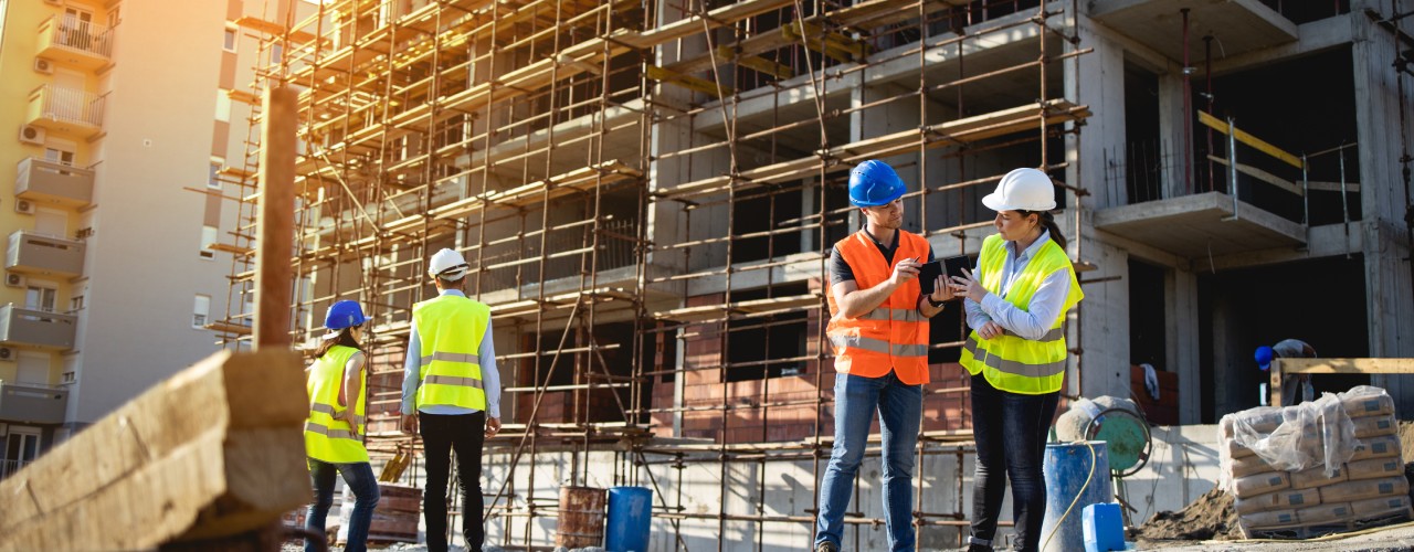 Four crew members stand on a construction site. All are wearing bright yellow and orange safety vests and hard hats.