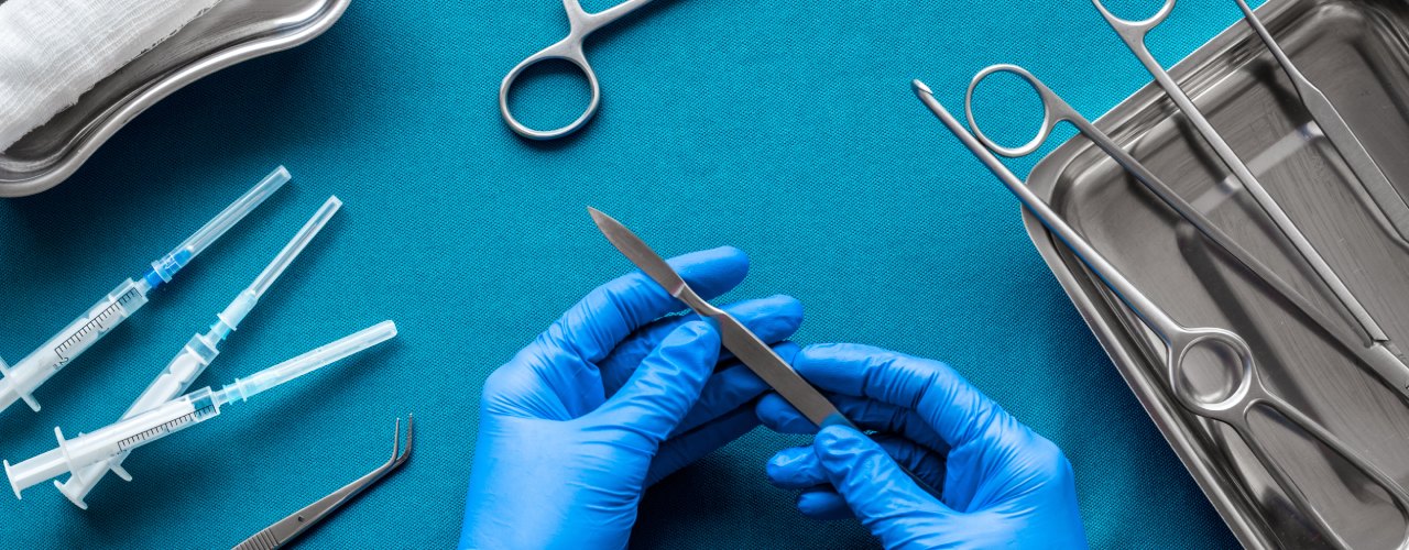 Doctor's hands holding a scalpel over a blue background with various surgical tools laid out on a table.