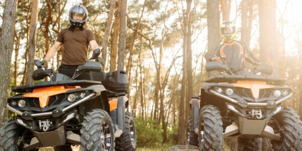 Two ATV riders wearing helmets and standing on their ATVs. The ATVs are parked on a dry, packed dirt trail in the woods.