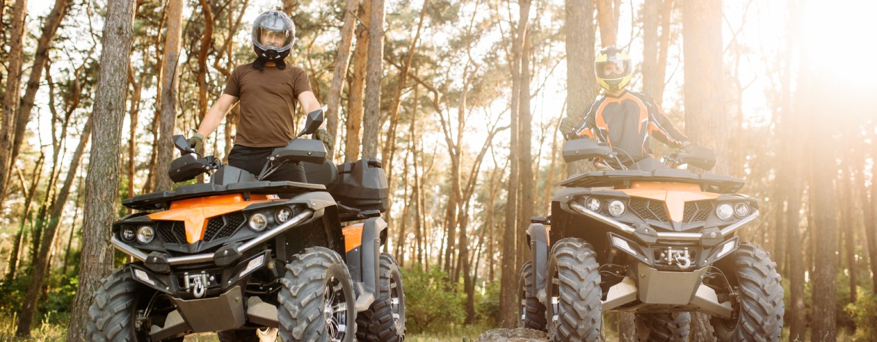 Two ATV riders wearing helmets and standing on their ATVs. The ATVs are parked on a dry, packed dirt trail in the woods.