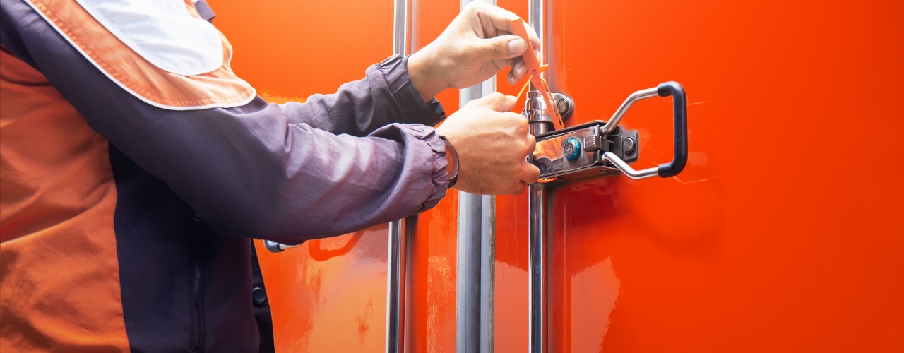 A delivery driver latches a security seal on the backside of a truck with an orange exterior to secure the shipment.