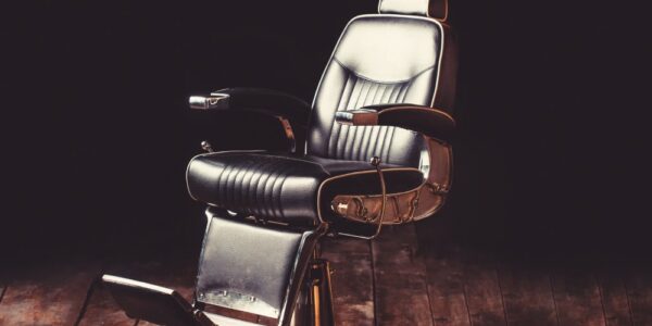 A single, old-school barber chair sitting in a spotlight on a worn-down wooden floor. Intense shadows fill the empty spaces.