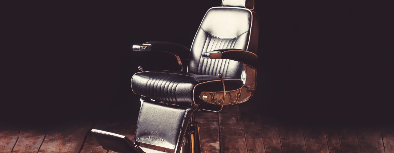 A single, old-school barber chair sitting in a spotlight on a worn-down wooden floor. Intense shadows fill the empty spaces.