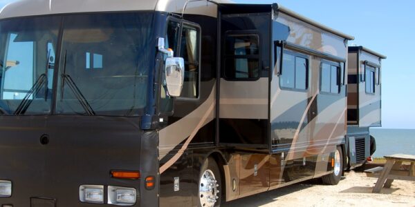 An RV in a parking lot next to a picnic table. The sky is clear and there is an ocean in the background.