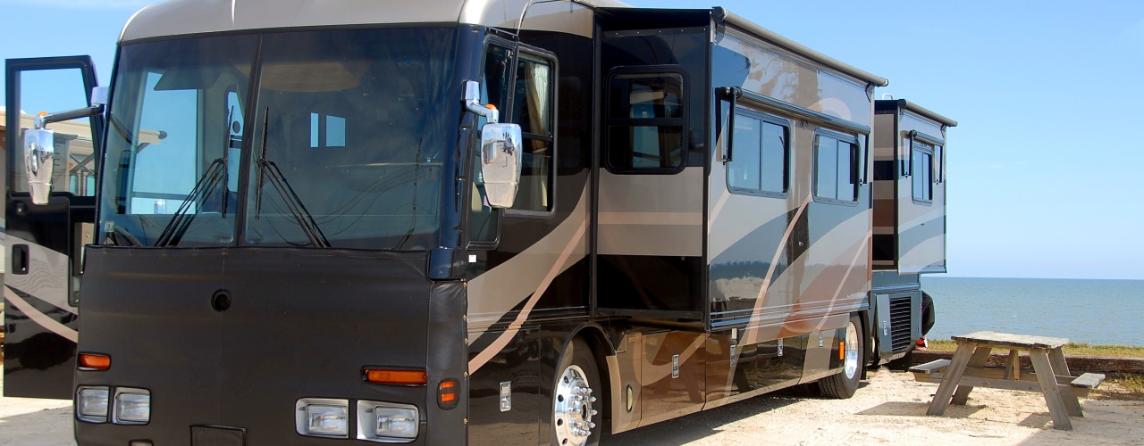 An RV in a parking lot next to a picnic table. The sky is clear and there is an ocean in the background.