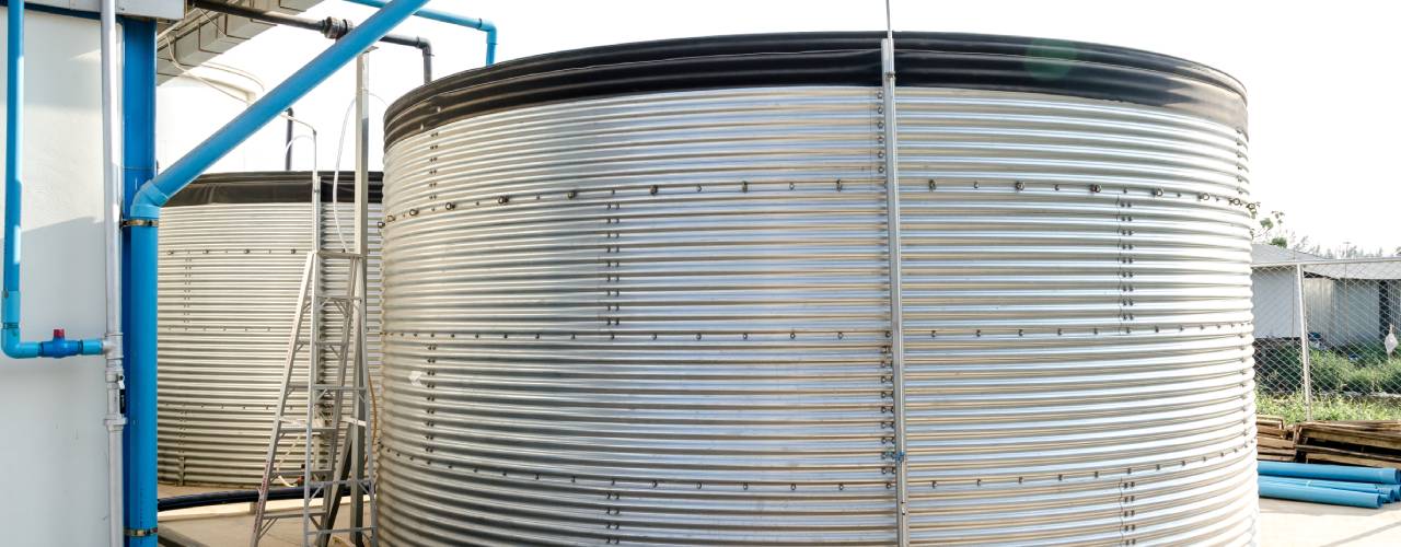A large ribbed silver holding tank with a black liner, connected by tubing to the side of a building.