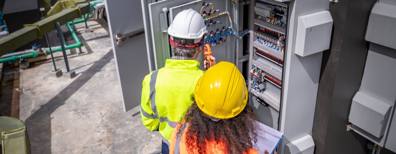 A male and female worker in hard hats and high-visibility vests inspect an electrical enclosure cabinet.