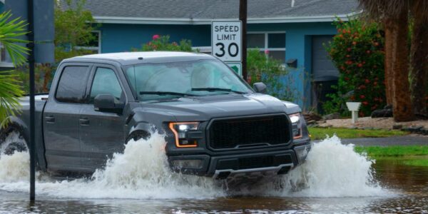 A black truck drives through a flooded residential street past a 30 MPH speed limit sign and blue house.