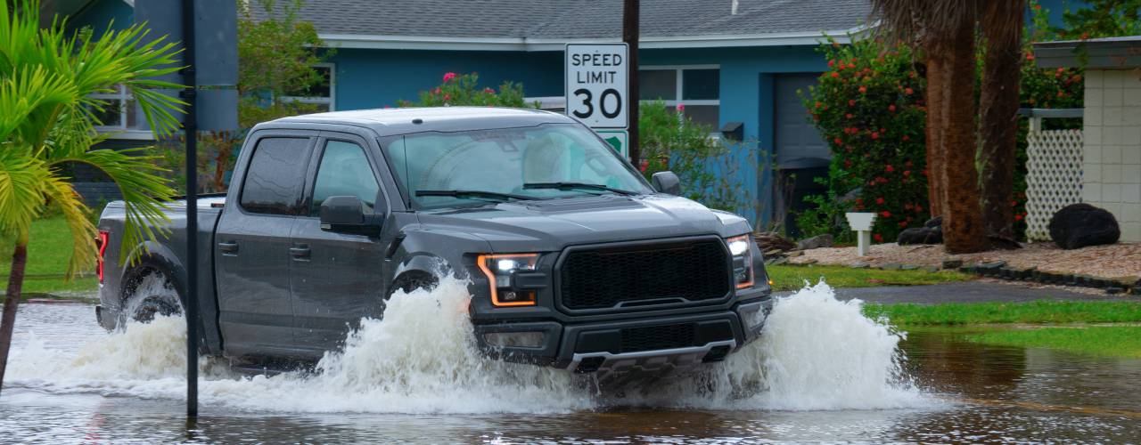 A black truck drives through a flooded residential street past a 30 MPH speed limit sign and blue house.