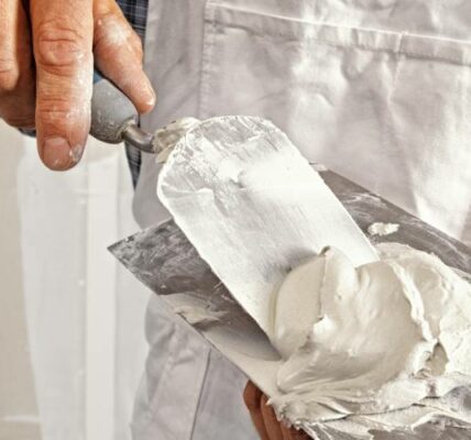 A man in white overalls and blue flannel scooping drywall onto a putty knife from a finishing trowel.