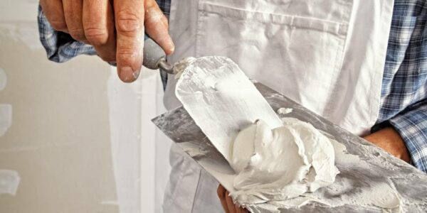 A man in white overalls and blue flannel scooping drywall onto a putty knife from a finishing trowel.