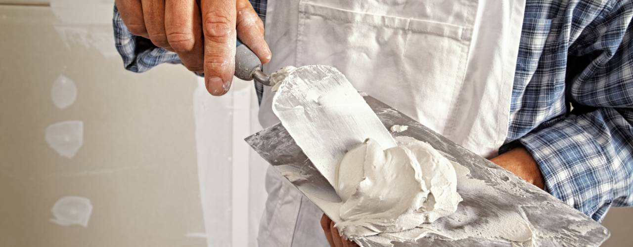 A man in white overalls and blue flannel scooping drywall onto a putty knife from a finishing trowel.