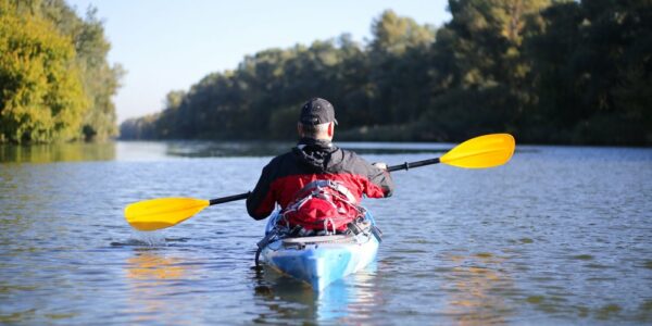 A man is sitting in a blue kayak and floating down a river lined with trees. He is holding his paddle above the water.