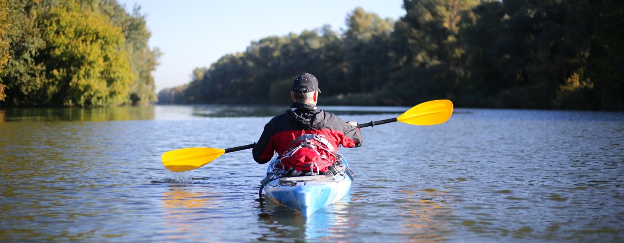 A man is sitting in a blue kayak and floating down a river lined with trees. He is holding his paddle above the water.