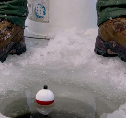 A close-up of an angler sitting on a white bucket next to a hole in the ice. They have a fishing line and a bobber in the water.