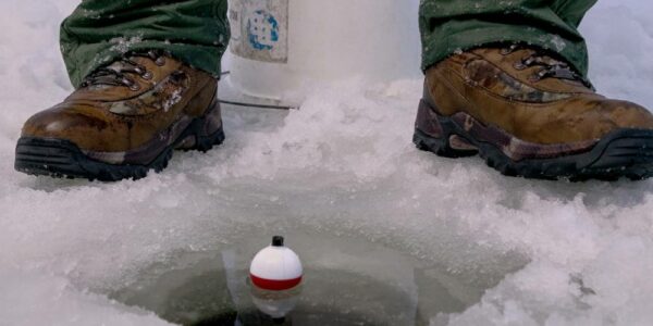 A close-up of an angler sitting on a white bucket next to a hole in the ice. They have a fishing line and a bobber in the water.