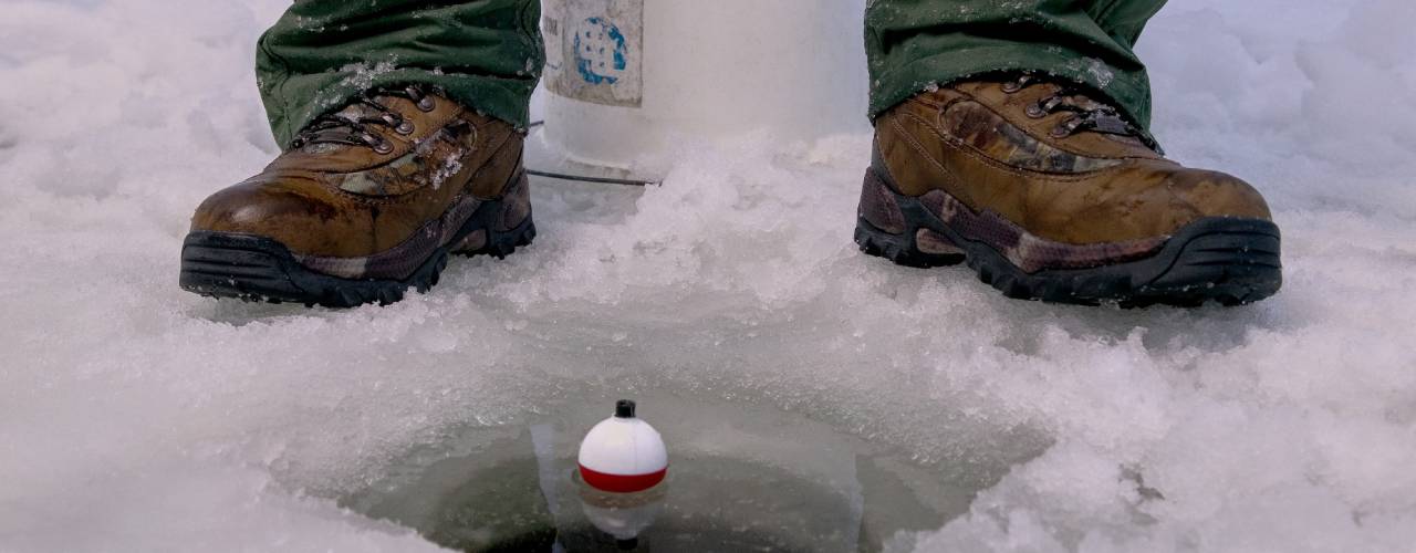 A close-up of an angler sitting on a white bucket next to a hole in the ice. They have a fishing line and a bobber in the water.