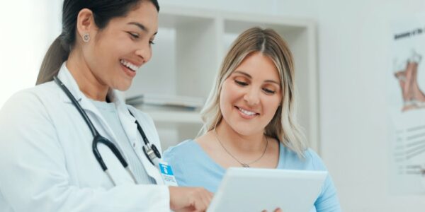 A pregnant mother sitting on the exam table, her delivery doctor is next to her with a clipboard. They're discussing a chart.