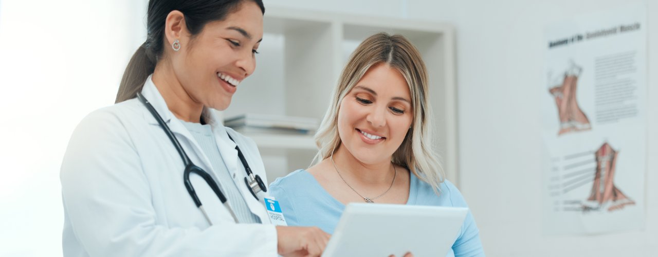 A pregnant mother sitting on the exam table, her delivery doctor is next to her with a clipboard. They're discussing a chart.