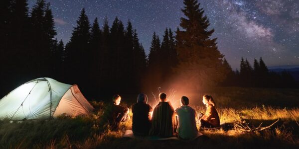 Five people are sitting on the ground at night next to a white tent surrounding a fire. The sky is full of stars.