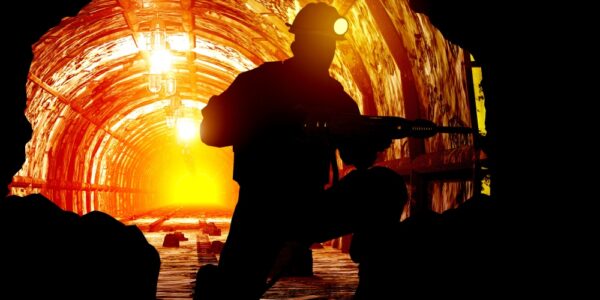 A miner wearing a helmet with a bright light on it is seen drilling in silhouette, in a mine, backed by orange light.