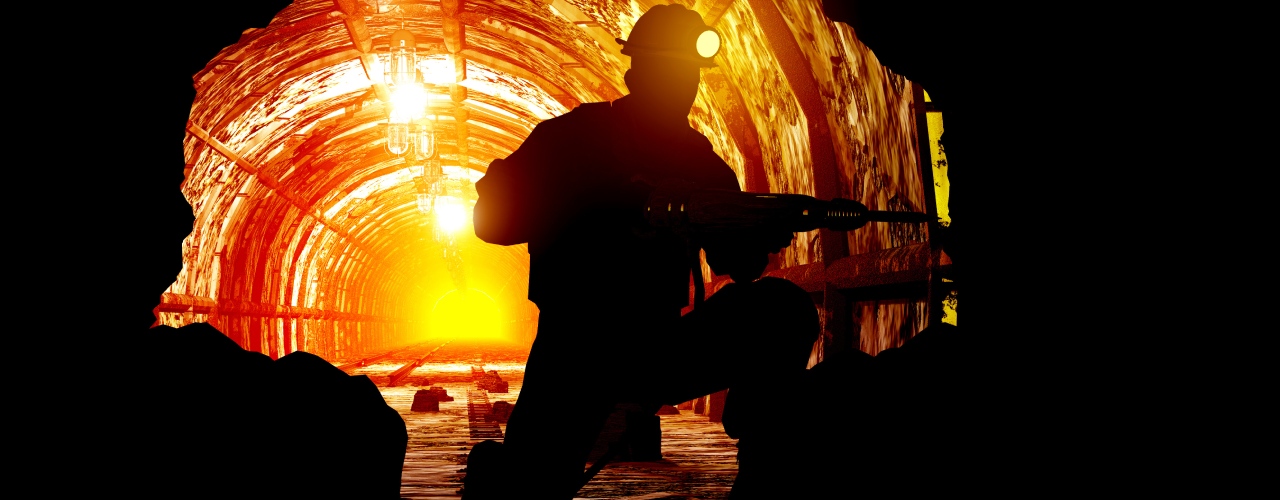 A miner wearing a helmet with a bright light on it is seen drilling in silhouette, in a mine, backed by orange light.