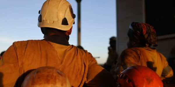 A group of miners covered in dirt sitting outside in the heat with their helmets on the table behind them.