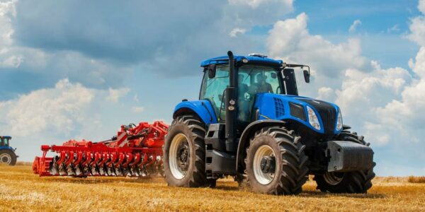 A blue tractor with a red harrow attachment cultivates a field with another tractor and the blue sky in the background.