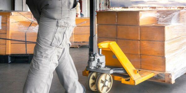 A worker using a pallet truck to move a small set of plastic-wrapped boxes out of a warehouse.
