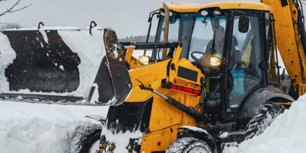 A yellow compact tractor using it’s front-mounted bucket to plow through large piles of snow, clearing paths in the land.