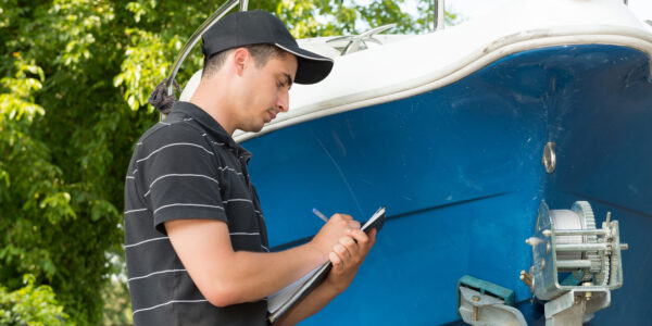 A man holds a notebook in front of a boat with a blue hull. He is writing something down with a pen.