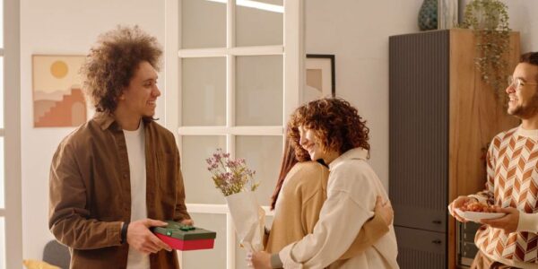 A man and woman couple welcome another couple into their kitchen. The guests bring flowers and a gift box.