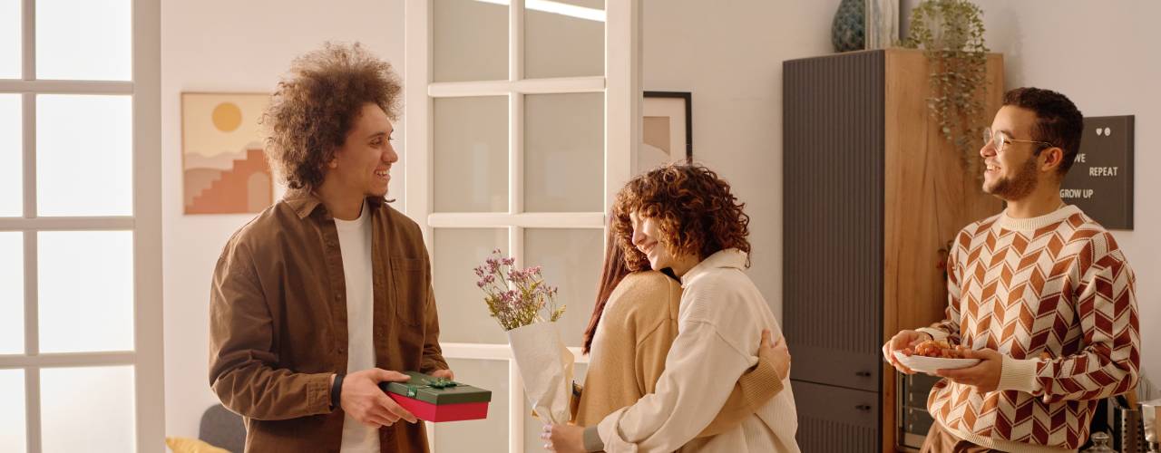 A man and woman couple welcome another couple into their kitchen. The guests bring flowers and a gift box.