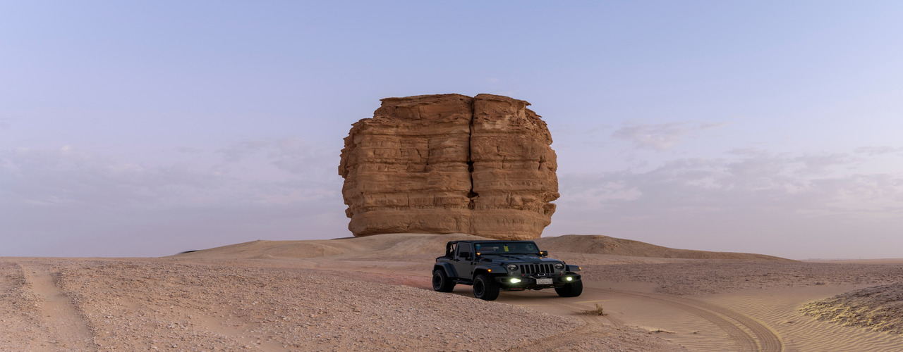A black jeep drives with its headlights on down an empty desert road near a large stone feature at dusk.
