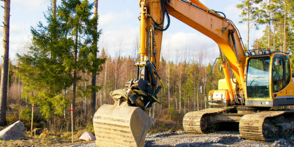 A yellow excavator sitting in the woods on a pile of rocks. The attachment is slightly bent and getting ready to grab rocks.