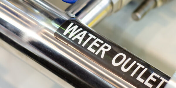 A close up of a section of stainless steel piping with a label that reads "water outlet" and blue valve handles attached.