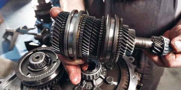 A top view of a mechanic in overalls, holding a gearbox with both hands in a brightly lit workshop.