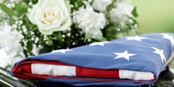 A close-up of an American flag folded into a triangle and resting on a black casket next to a bouquet of white roses.