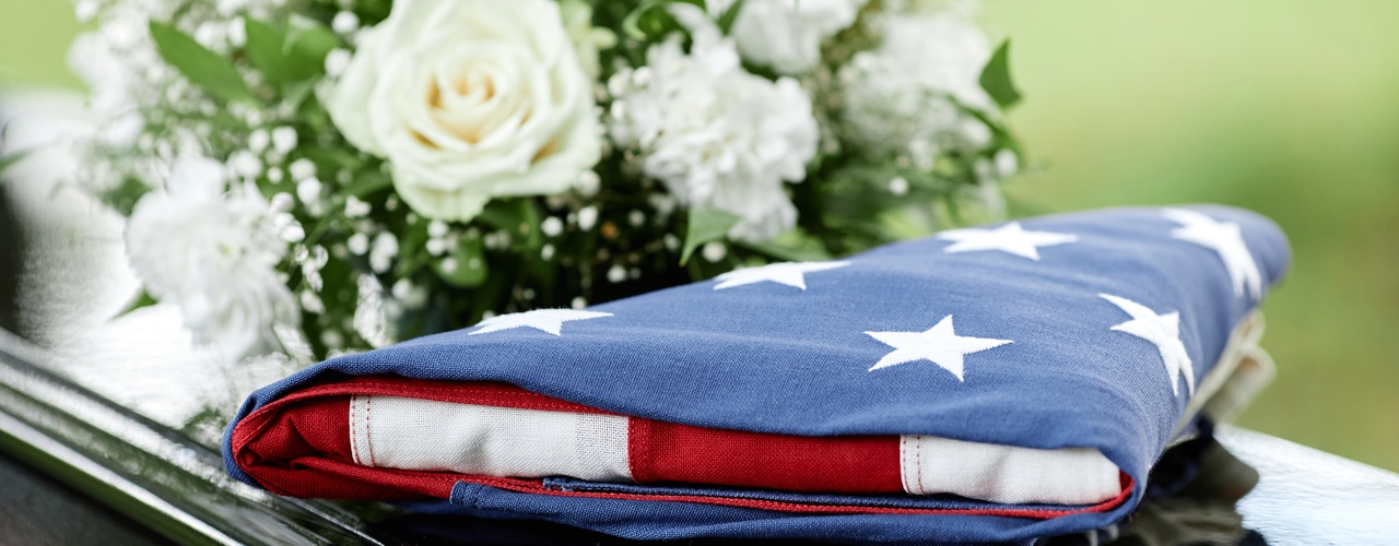 A close-up of an American flag folded into a triangle and resting on a black casket next to a bouquet of white roses.